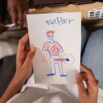 A high-angle view of a child's drawing of their father, placed in front of a toy house, showing family creativity.